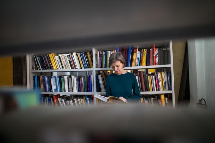 Woman browsing books in a library as viewed through a space in an adjacent bookshelf.